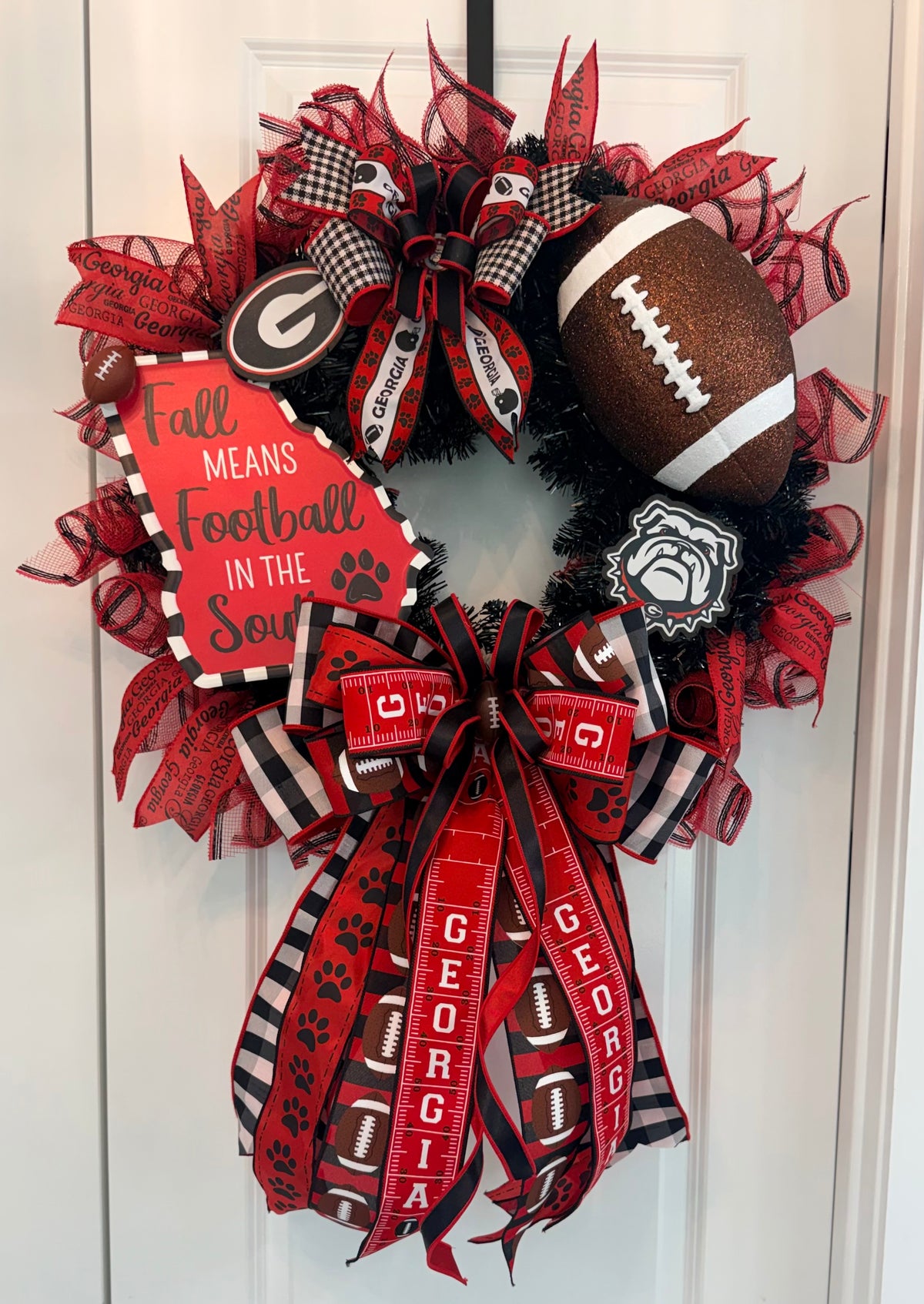 A Georgia Bulldogs themed fall wreath hanging on a white door. The wreath features red and black mesh ribbons, a football, a sign that reads 'Fall Means Football in the Soul', and a large bow with 'GEORGIA' printed on it.